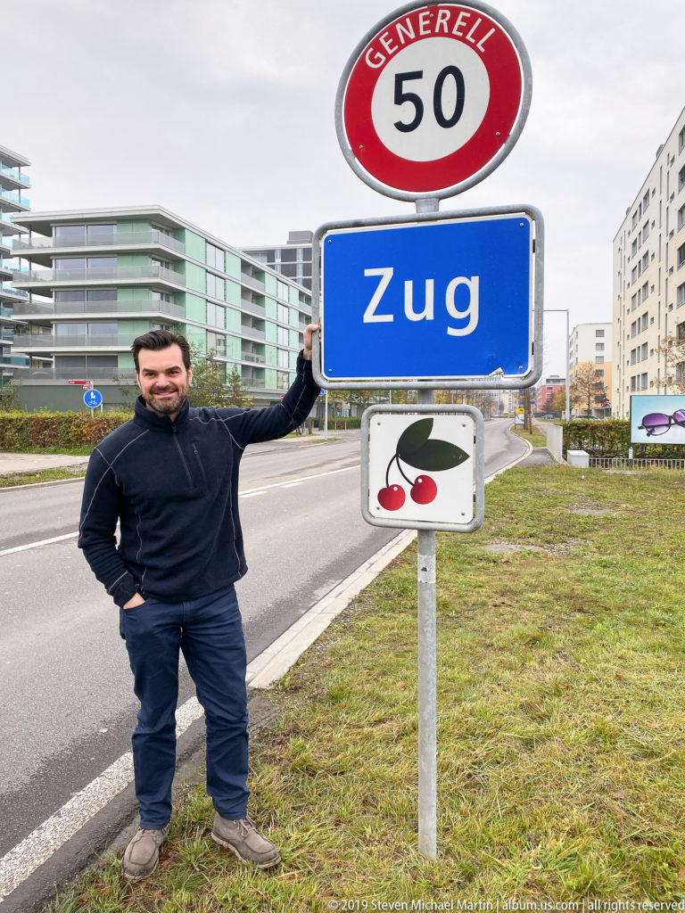 Man standing beside a blue Zug road sign beneath a 50 km/h speed-limit sign and cherry emblem.