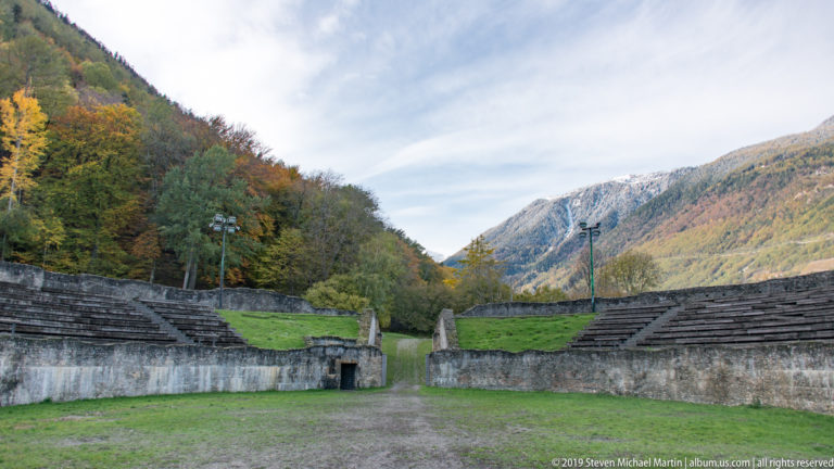 View from a grassy amphitheatre arena toward stepped seating, a central passage, autumn trees, and Alpine mountains.