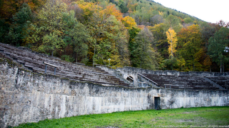 Curving stone seating and high arena wall of an amphitheatre backed by colorful autumn woodland.