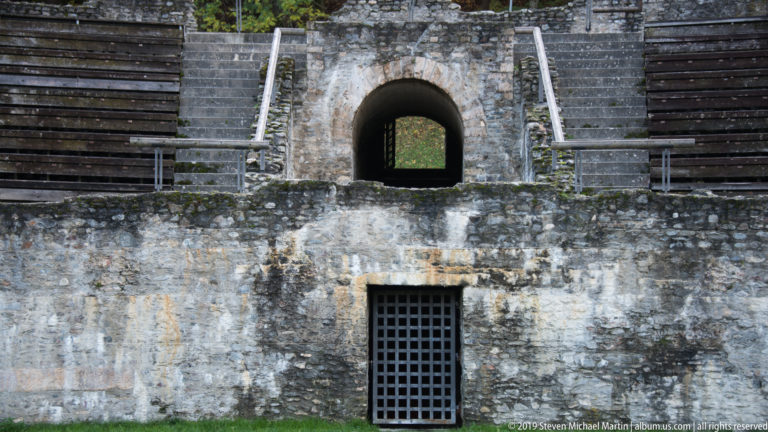 Stone amphitheatre wall with an upper arched passage, lower barred opening, and stepped seating on both sides.