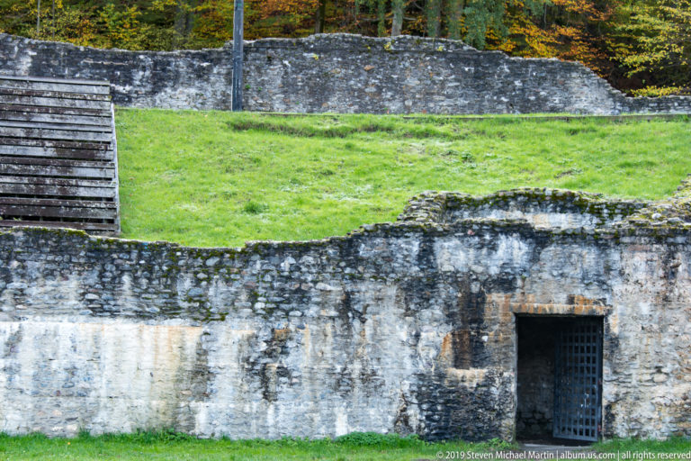 Weathered amphitheatre wall with a small barred doorway below a grassy embankment and stone retaining wall.