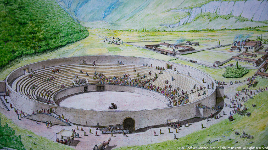 Illustrated reconstruction of an elliptical Roman amphitheatre filled with spectators in a broad Alpine valley.