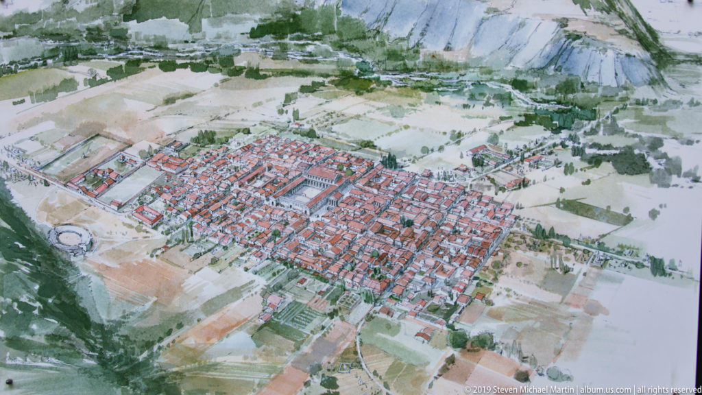 Bird’s-eye painted reconstruction of a Roman town grid surrounded by fields and steep mountain slopes.