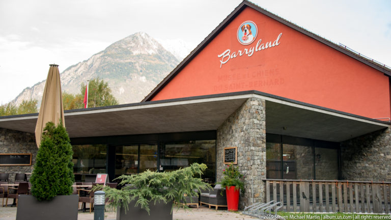 Red-gabled Barryland building with logo, stone entrance columns, terrace seating, and an alpine mountain behind it.