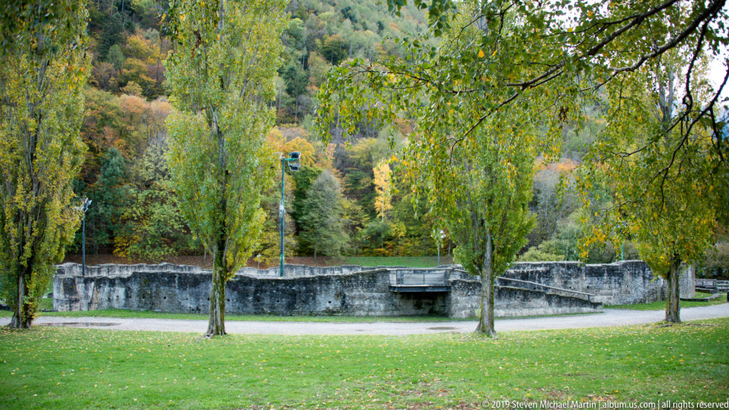 Low ancient stone walls in a grassy, tree-filled archaeological park with wooded hills in the background.