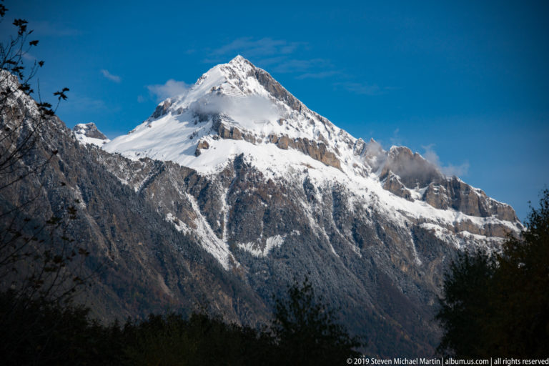 Snow-covered Alpine summit above shadowed rocky ridges near Martigny, framed by dark foreground branches beneath a deep blue sky.