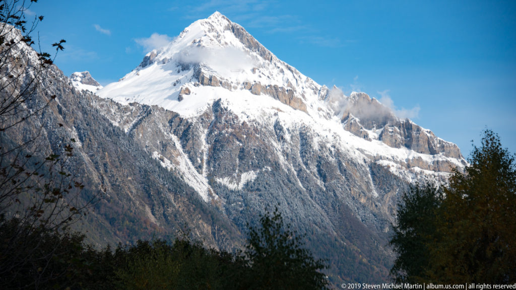 Bright snow-covered pyramidal Alpine peak rises above dark rocky ridges near Martigny under a clear blue sky with wisps of cloud.