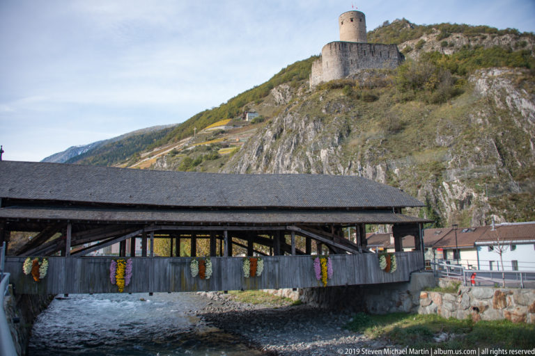 Decorated covered wooden bridge in the foreground with a round-towered stone castle on the rocky hillside above.