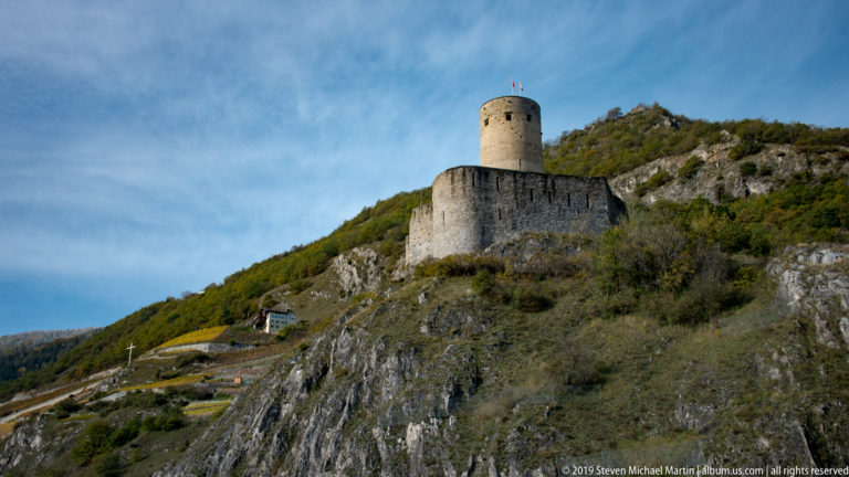 Round stone castle tower and curtain wall on a rocky, shrub-covered hillside beneath a blue sky.