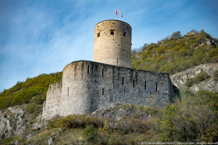 Close view of a round stone castle keep with flags above a thick curtain wall on a scrub-covered rocky slope.