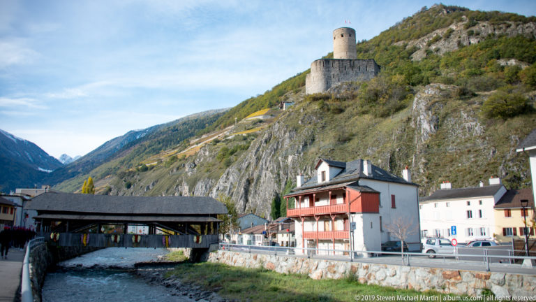 Covered wooden bridge over a river with houses along the bank and a round-towered castle high on the rocky hillside.