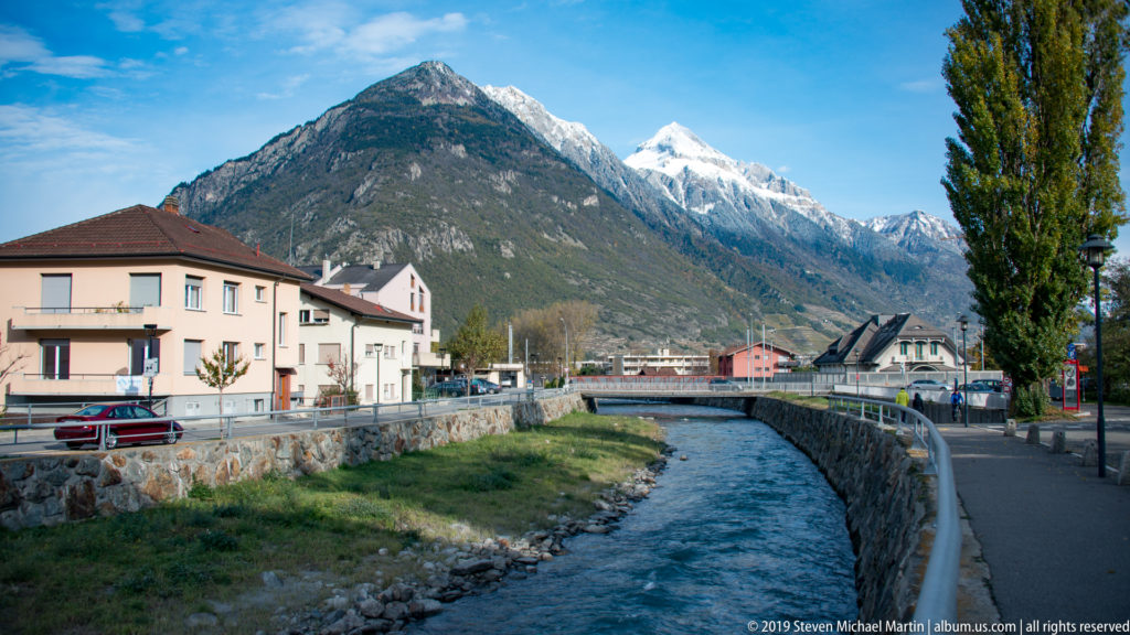 Stone-lined river channel through town with houses, road, bridge, tall trees, and Alpine mountains ahead.