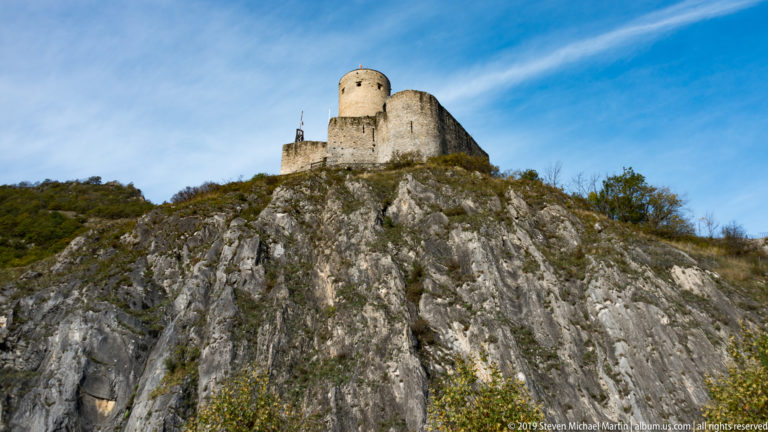Stone castle with cylindrical keep perched high on a rugged gray rock outcrop beneath a blue sky.