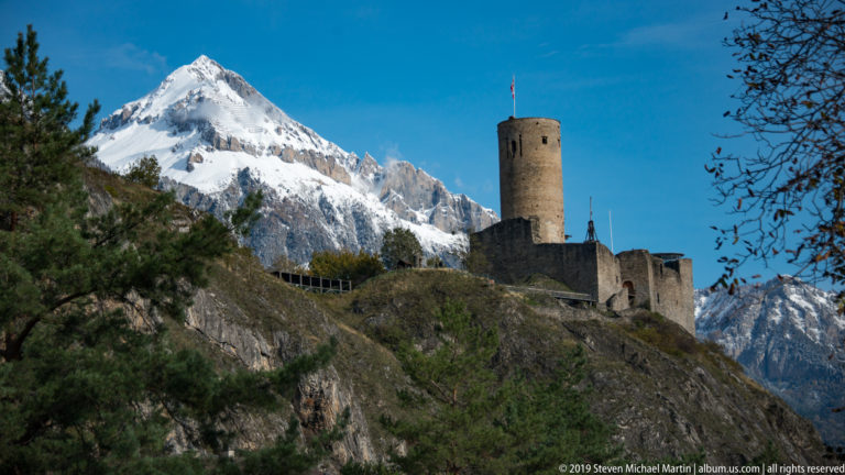 Distant stone castle with round tower on a wooded rocky ridge, with a snow-covered mountain rising behind.