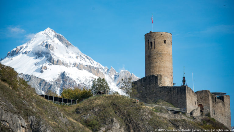 Round stone castle keep and lower walls on a rocky slope, backed by a large snow-covered Alpine peak.