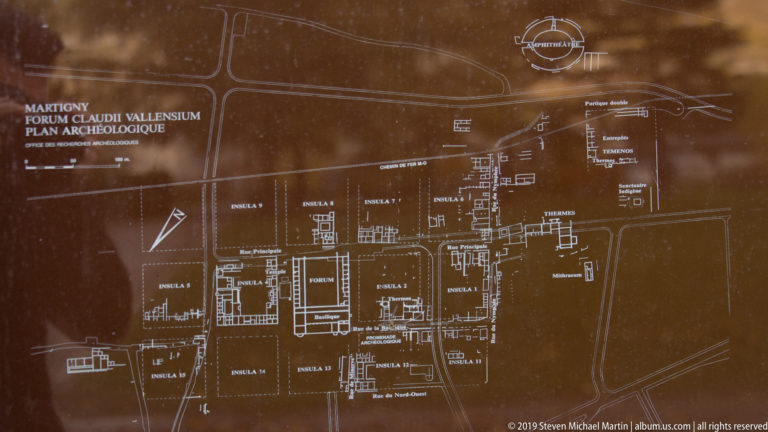 Brown archaeological map labeled Forum Claudii Vallensium, with insulae, streets, structures, and amphitheatre marked.