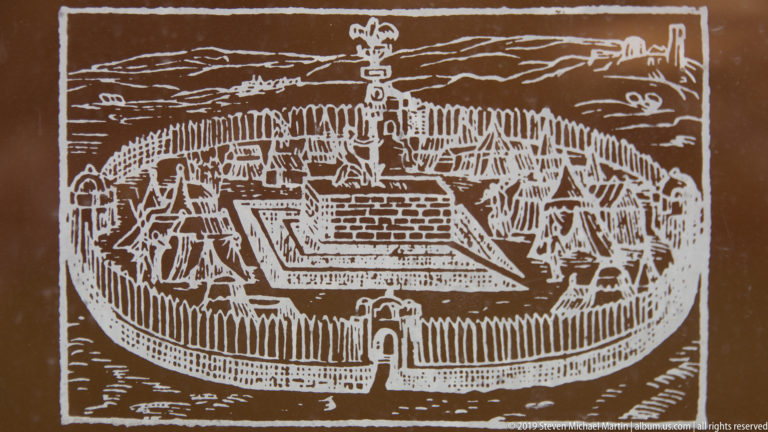 White line drawing on brown background showing a compact fortified enclosure with buildings, perimeter defenses, and a central tower.