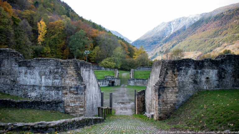 Stone-walled entrance leading into an amphitheatre arena, with grassy banks, autumn trees, and mountains beyond.