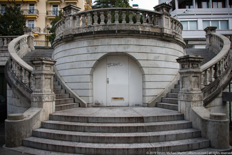 Symmetrical curving stone staircases rising around a rounded central terrace with a recessed white doorway.