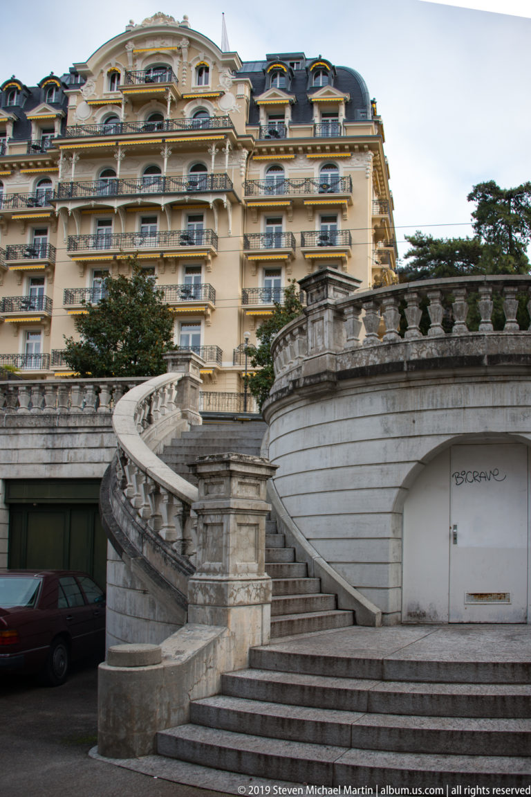Broad curving stone staircase and balustrades rising toward a grand cream-colored hotel with mansard roof.