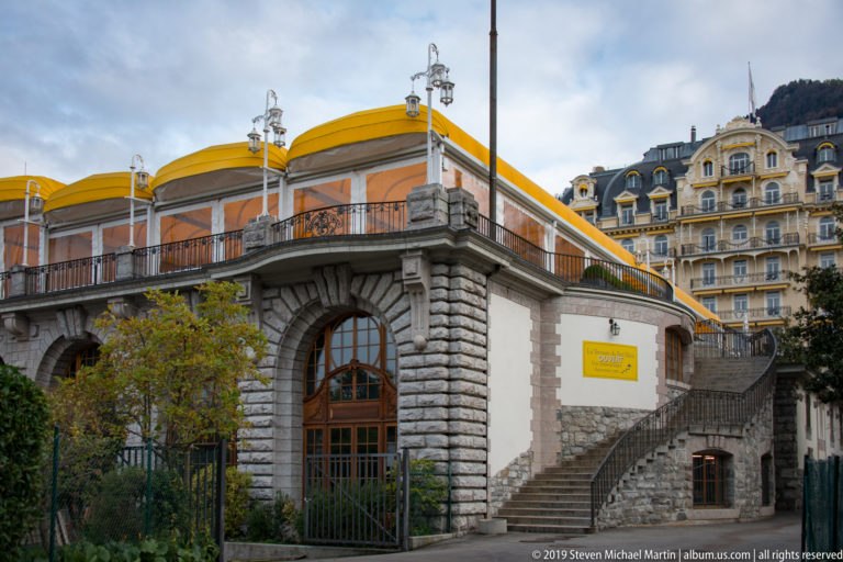 Historic stone building with tall arched entrance, balustraded terrace, exterior stairs, and bright yellow roofline in Montreux.