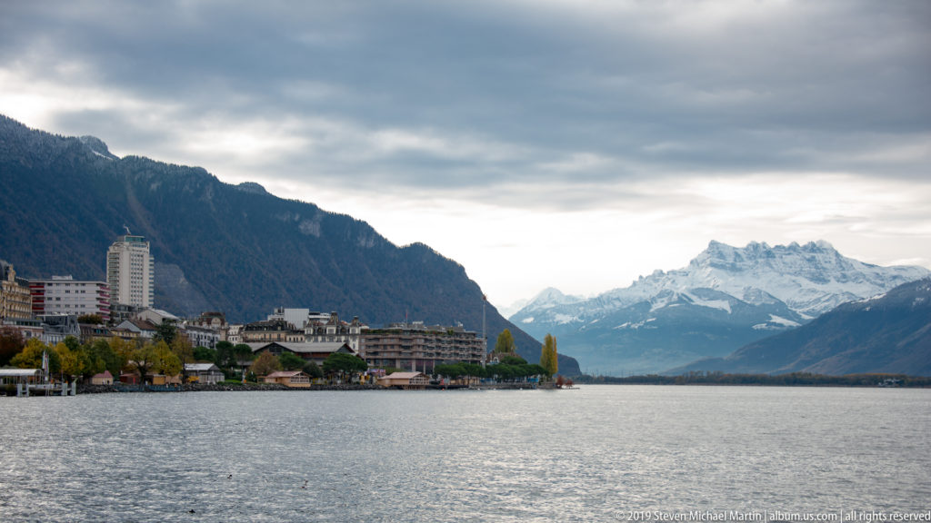 Montreux shoreline across Lake Geneva beneath a steep mountain, with snow-covered peaks in the distance.