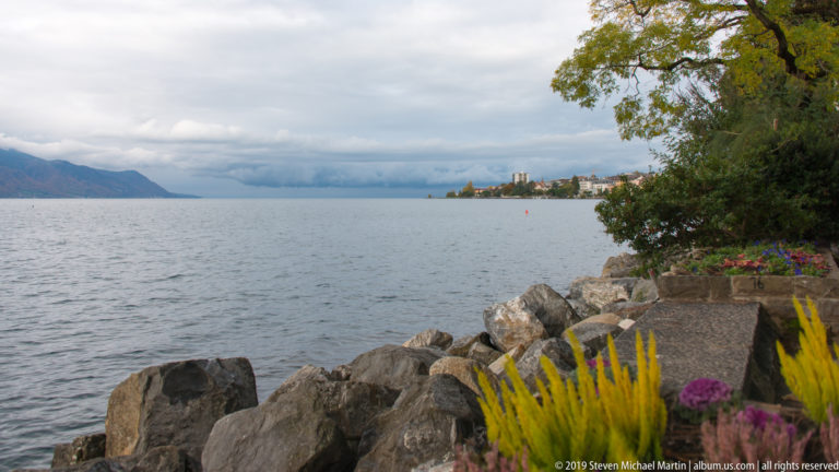 Large shoreline rocks and yellow garden plants beside Lake Geneva, with distant wooded slopes beneath cloudy sky.
