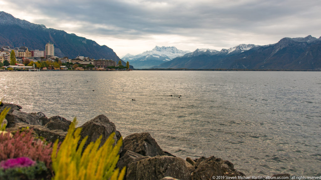 Rocky landscaped Lake Geneva shore with Montreux at left and snow-dusted mountains extending into the distance.