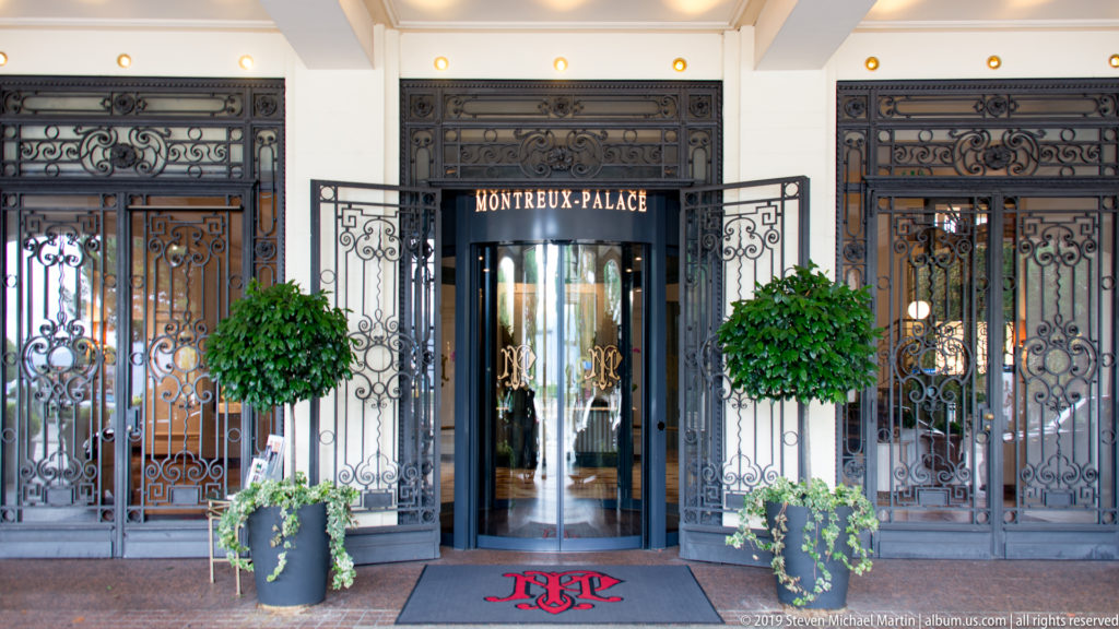 Ornate wrought-iron and glass entrance marked “MONTREUX-PALACE,” flanked by two clipped potted trees.