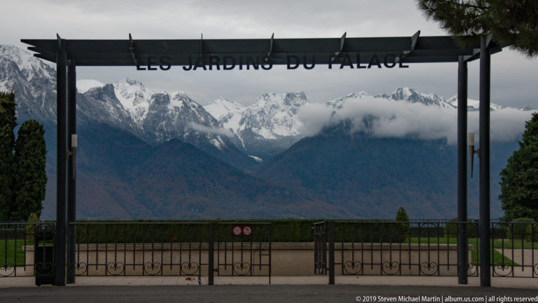 Gateway labeled “LES JARDINS DU PALACE” framing clipped gardens and cloud-wrapped Alpine mountains beyond.