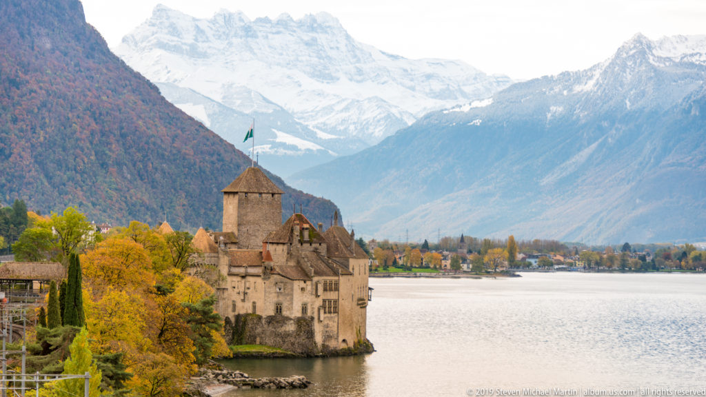 Château de Chillon beside Lake Geneva with autumn trees and snow-dusted Alpine mountains rising behind.
