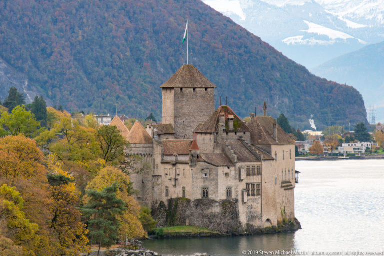 Château de Chillon framed by yellow autumn trees beside Lake Geneva with mountains behind.