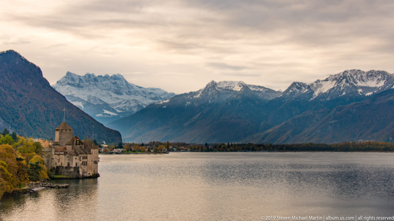 Wide Lake Geneva view with Château de Chillon at left and snow-covered Alpine peaks beyond the water.