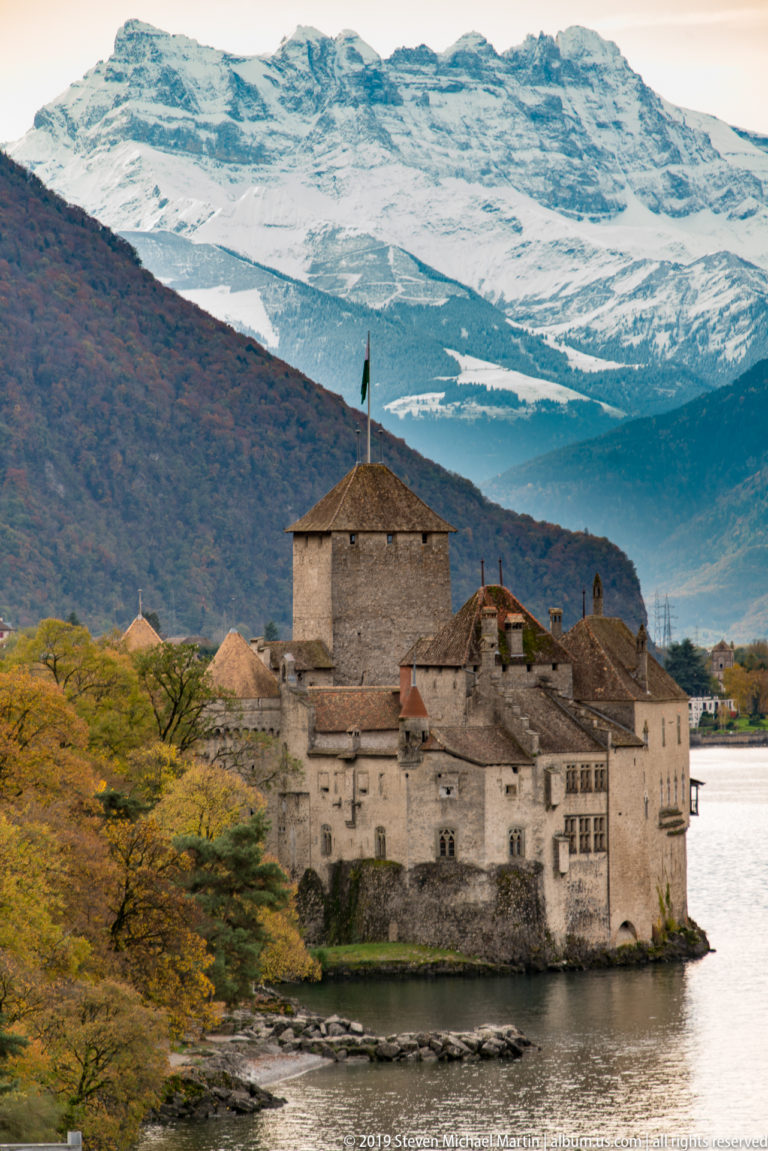 Château de Chillon projecting into Lake Geneva beneath broad snow-covered Alpine peaks and autumn trees.
