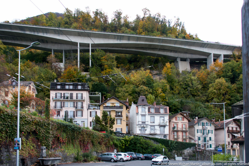 Colorful hillside residences, parked cars and overhead wires beneath a large elevated concrete structure in Montreux.