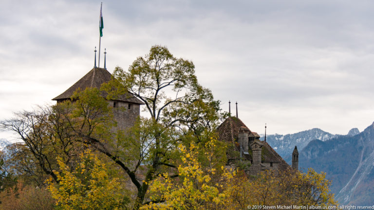 Stone towers and steep roofs of Château de Chillon rising through autumn trees with mountains behind.