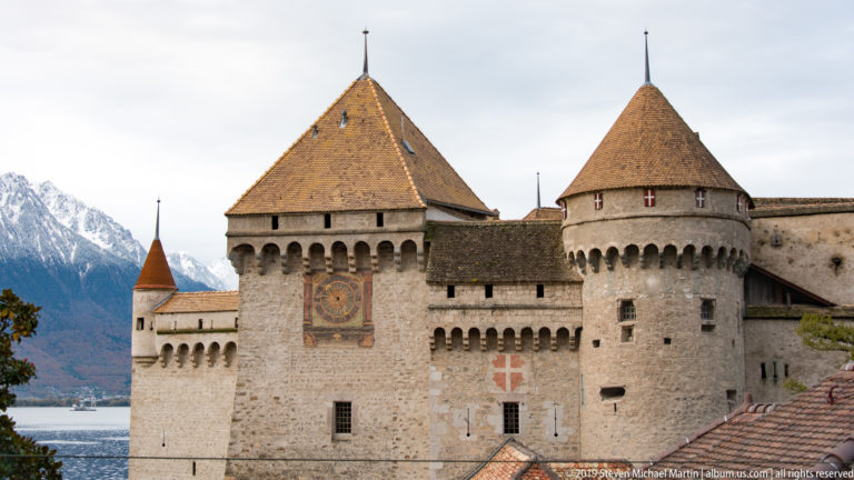 Fortified stone façade of Château de Chillon with clock tower, round tower, tiled roofs, and mountains at left.