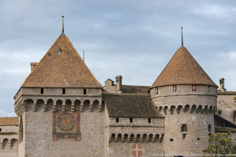 Square castle tower with large decorative clock face beside a round conical-roofed tower at Château de Chillon.