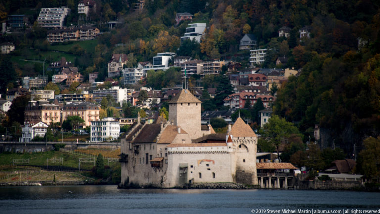 Château de Chillon on the shore of Lake Geneva with hillside homes and autumn woodland rising behind.