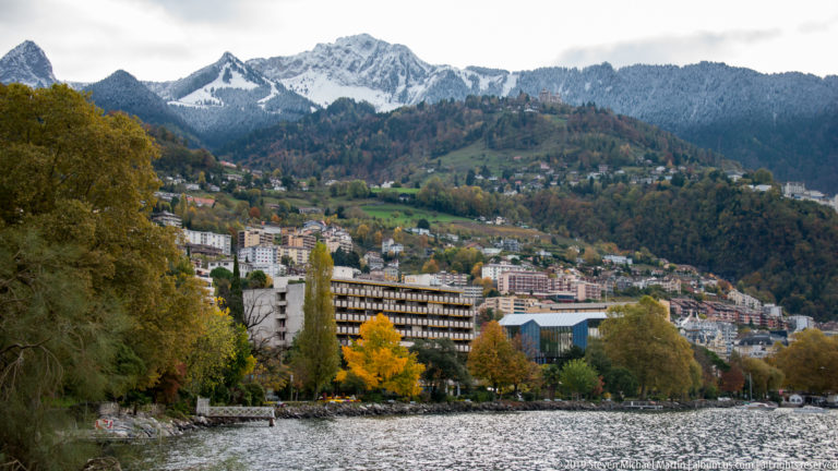 Montreux buildings and autumn trees climbing a steep hillside above Lake Geneva with snowy ridges behind.