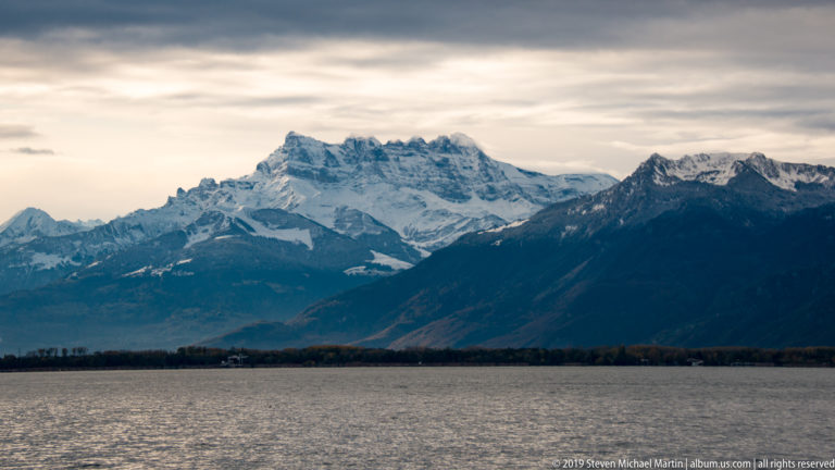 Snow-covered Alpine massif rising across Lake Geneva beneath a softly lit overcast sky.