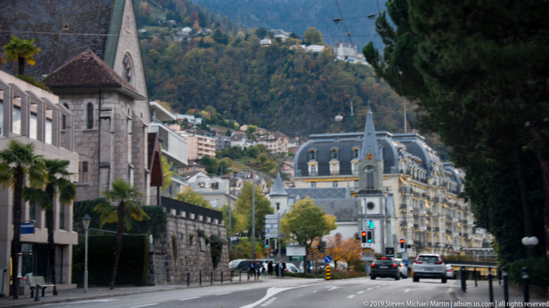 Curving Montreux avenue with church, ornate mansard-roofed buildings, cars, palms, and hillside development.