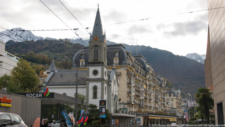 Montreux street with slender church tower, ornate hotel façades, traffic, overhead wires, and wooded mountain behind.