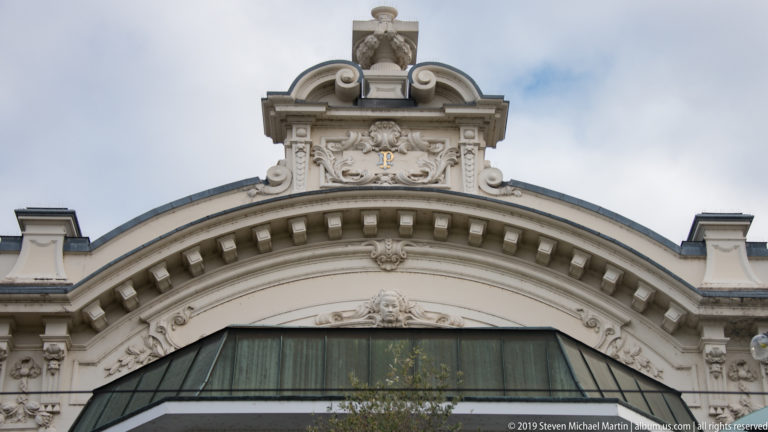 Ornate pale arched façade with carved scrolls, masks, relief figures, curved cornice, and a small crowning sculpture.