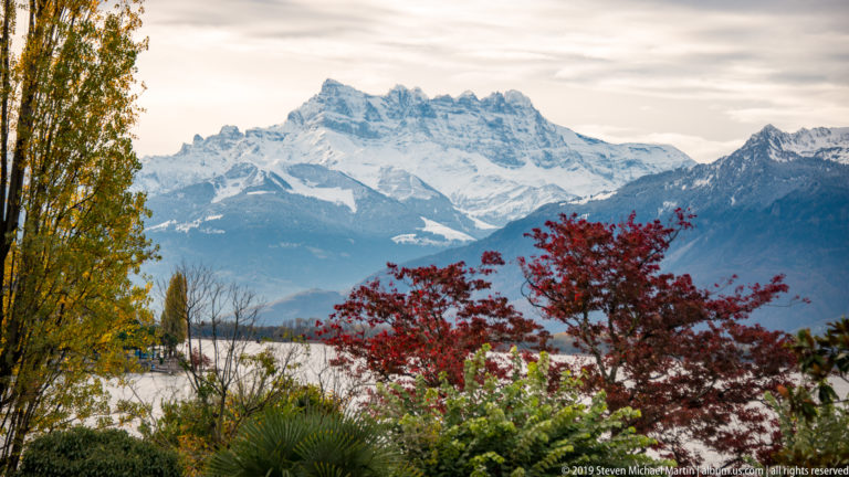 Snow-covered Alpine mountains beyond Lake Geneva, framed by red and yellow autumn trees, palms, and garden shrubs.