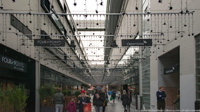 Covered Zurich pedestrian passage with shoppers beneath rows of suspended lights and signs for Four Points and Crédit Suisse.