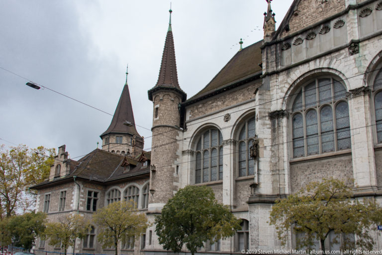 Closer view of Swiss National Museum wall with tall windows, a round turret under a conical roof, and trees.