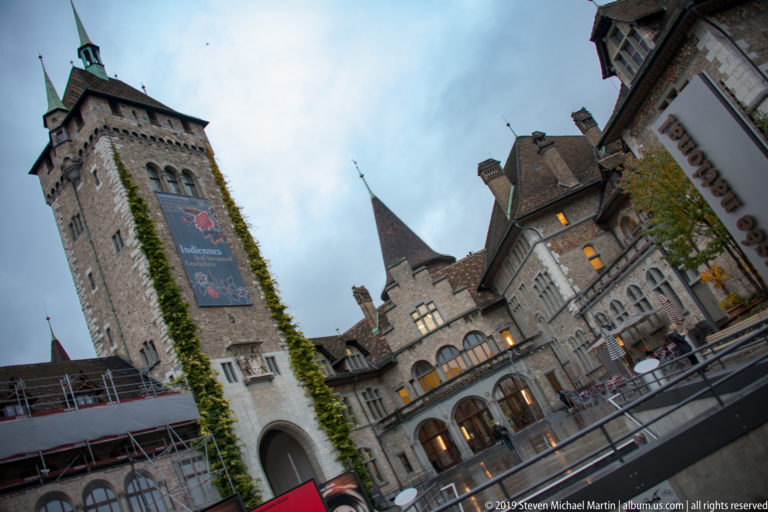 Tilted view of Swiss National Museum tower, ivy-covered stone walls, arched gateway, steep roofs, and scaffolding.