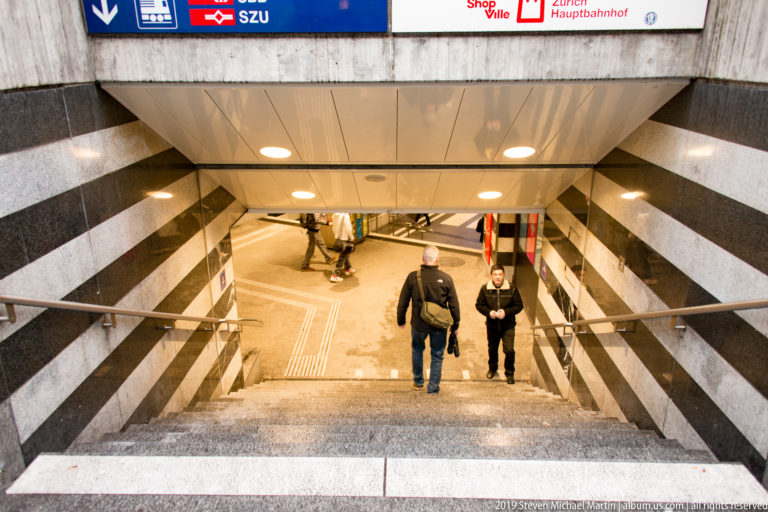 People descend wide stairs into Zürich Hauptbahnhof beneath S-Bahn signs, striped stone walls, and metal handrails.