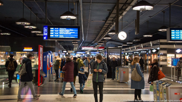 Travelers in Zürich Hauptbahnhof concourse beneath platform signs, departure boards, clocks, lights, and retail kiosks.